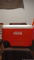 Side view of Coca-Cola Igloo wheelie cooler showing red body, white lid, black wheels, and handle.
