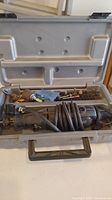 Opened grey plastic carrying case showing Dremel rotary tool with power cord coiled inside and assorted accessories fitted into compartments.