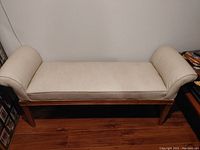 Front view of an off-white vinyl cushioned bench with wooden frame and fluted wooden legs, curved padded armrests on each end.