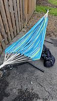 Hammock fabric in blue and green stripes laid on the metal stand with carrying bag beside it on asphalt ground near wooden fence.
