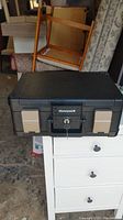 Closed black Honeywell safe with beige side latches and key lock on white drawer unit, showing front and top angles.