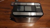 Bread maker exterior showing stainless steel finish, digital control panel on right, viewing windows on top lid, Kuradori logo visible