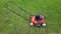 Side view of red Yardmachines lawn mower on grass showing black handle and four white wheels.