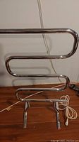 Front view of Warmrails chrome towel warmer showing three bars, base, and power cord on wooden floor.