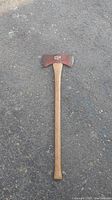 Full view of the double bit axe showing the wooden handle and red painted metal axe head laying on asphalt.