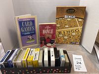Photo showing 14 decks of playing cards in plastic boxes, two card game books, and a bingo game box arranged in a tray.