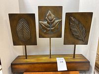 Photo showing three metal leaf designs on rectangular plates mounted on metal rods attached to a wooden base. The centerpiece is used and may need slight tightening of screws.