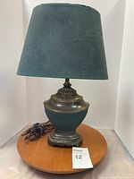 Green lamp with fabric shade showing front view, the lamp base is green with bronze/brass accents and round shape base with visible power cord.