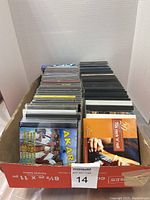 Top view of box filled with stacked used music CDs in jewel cases and some cassette tapes, showing album covers and titles on spines.