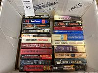 Photo shows a large box filled with various used novels arranged in two rows. Titles include works by Tom Clancy, Dean Koontz, Michael Crichton, Carl Sagan, Robert Ludlum, and others.
