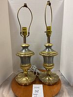 Pair of silver and gold decorative table lamps on a wooden surface with white electrical cords, no shades, with harp holders visible.