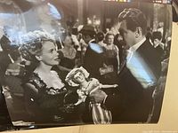 Photo of a black-and-white 8 x 10 movie still showing Angela Lansbury and a male actor in 19th-century costume holding a doll in a crowded ballroom setting.