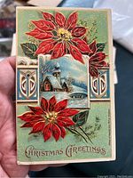 Front of a Christmas postcard featuring bright red poinsettia flowers, a snowy winter church scene, and decorative gold accents with the text 'Christmas Greetings'.