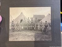 Cabinet photo showing group of New Hampshire National Guard officers seated outdoors in front of tents and wooden building, dated 1909
