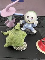 Four Disney Nightmare Before Christmas cast figurines displayed on a black surface, including a purple witch, a blue-suited skeleton figure, a green monster with a chip, and part of a red and cream figurine.