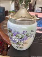 Side view of white porcelain biscuit jar with vivid hand-painted violet flowers and green leaves, silver plated lid and handle visible.