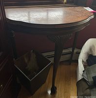 Side view of the wooden demilune table with claw feet and brown waste bin placed beneath the table, showing wear and peeling veneer.