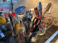 Photo showing oversized glass beer mug with red text, kitchen utensils including scissors, and stainless steel flatware in containers on kitchen counter.