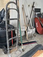 View of hand truck, push broom, shovel, rakes, and red wheelbarrow with stacked tires and other items in background