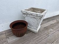 Wide view showing both the large square concrete outdoor planter and the smaller round metal planter positioned next to each other on an outdoor floor against a white wall.