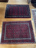 Two red rectangular area rugs on wood floor, seen from above. Both rugs show intricate traditional geometric and floral patterns with no fringes at edges.