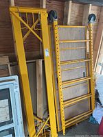 Yellow steel vertical scaffolding frames with assembly holes leaning against wood wall inside garage.