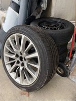 Four tires stacked, one tire with a silver multi-spoke alloy rim in front showing the tread and overall weathered condition.