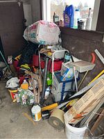 Wide-angle photo of assorted yard and garden tools, hardware, containers, and other items stacked in a corner of a garage or storage area.
