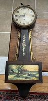 Full view of banjo clock showing the wooden case, floral decoration, and scenic panel.