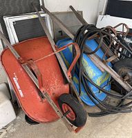 Photo showing two wheelbarrows leaning against a wall, one orange with 'Tote Temper USA' branding, the other blue with wooden handles, and a black garden hose coiled on top.
