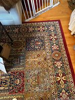 Photo showing a corner of the rug near furniture, highlighting the multicolored floral block patterns and the wide red border.