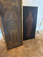 Two medieval figure art pieces side by side, one framed under glass and the other unframed on carpeted floor.