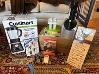 Full lot contents arranged on patterned rug showing blender box, salt and pepper shaker box, popsicle molds, Paula Deen disc set, flour sack towel, Yonanas blender