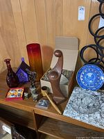 Wide shot showing grouped collection of decorative items including vases, birdhouse, figurines, plate, tiles, wood sculptures, and gavel on wooden shelves.