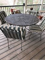 Full view showing round iron mesh table with umbrella hole and four matching iron chairs with striped seat and back cushions on wooden deck porch.