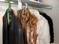 Four women's outerwear items on hangers: one black leather jacket, one brown fur vest, one white fur short jacket, one black fur short jacket.