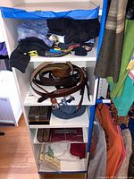 Three-shelf white organizer showing folded medium T-shirts on top, four leather belts on middle shelf, blue fabric bin and maroon Diplomat folder on bottom.