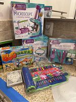 Lot overview showing all items on kitchen counter including boxed microscope kit, journals, bug lab container, gel pens set, bug catcher, and board game.
