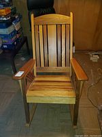 Front view of wooden rocking chair with vertical slats and flat armrests on a tiled floor in a basement room, cushions not visible.