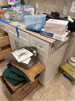 Photo showing several boxes of Rodan + Fields Proactiv beauty supplies, a blue storage container, and a men's grooming set with black carrying bags on the countertop. Below countertop a box is filled with tan and dark green towels.