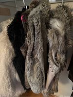 All outerwear hanging on a rack, displaying four distinct vests made of faux fur or shearling materials in various colors.