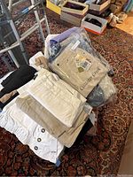 Stack of women's pants and tops with some pants in neutral colors including pants with tags showing Coldwater Creek brand.