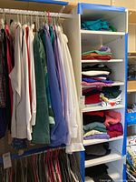 Hanging extra large women's shirts, sweaters, blouses in various colors on hangers next to a tall shelving unit with folded clothes stacked on shelves.