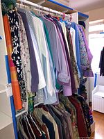 Two-level clothing rack with various women's blouses, tops, slacks, and vests on hangers. Includes floral and solid colors with pastel hues like lavender and white.