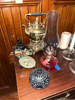 Photo showing the brass chafing kettle, red glass oil lamp with chimney, mosaic elephant, stone squirrel, and enameled plate with stand on wooden surface.
