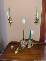 Two brass wall-mounted candle sconces with white taper candles hung on the wall above a wooden surface that holds two decorative brass candleholders with faceted glass pendants and green candles, and one additional brass candleholder.