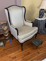 Full view of vintage wingback armchair with matching footstool on hardwood floor near lamp and side table.