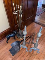 Side view of brass fireplace tools in stand, brass andirons, and black cast iron dog door stop placed on wooden floor.
