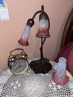 Tulip lamp with two shades, extra glass shade, and brass twin-bell alarm clock on white crocheted tablecloth.