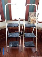 Front view of two folding step ladders with white frames and black steps, showing paint splatter wear.